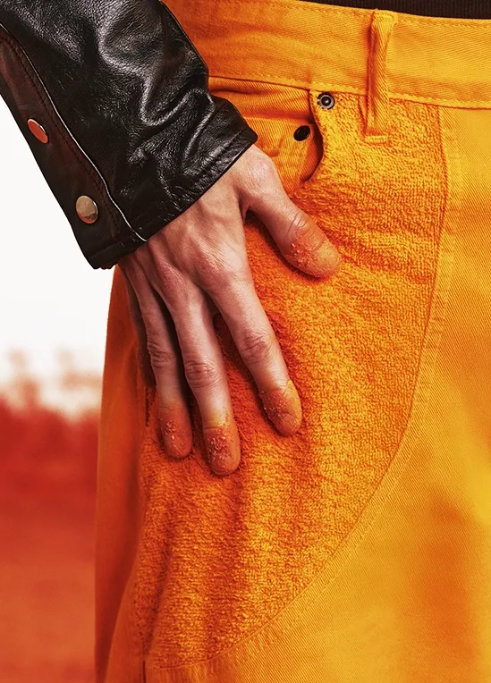 A stylish young man with curly hair sits on an orange block, wearing a black leather jacket, black turtleneck, bright orange pants, and matching sneakers. He sports futuristic orange sunglasses and holds a bag of Cheetos Crunchy in one hand while eating a chip with the other. The background is a white-to-orange gradient, resembling a fiery Cheetos dust explosion.